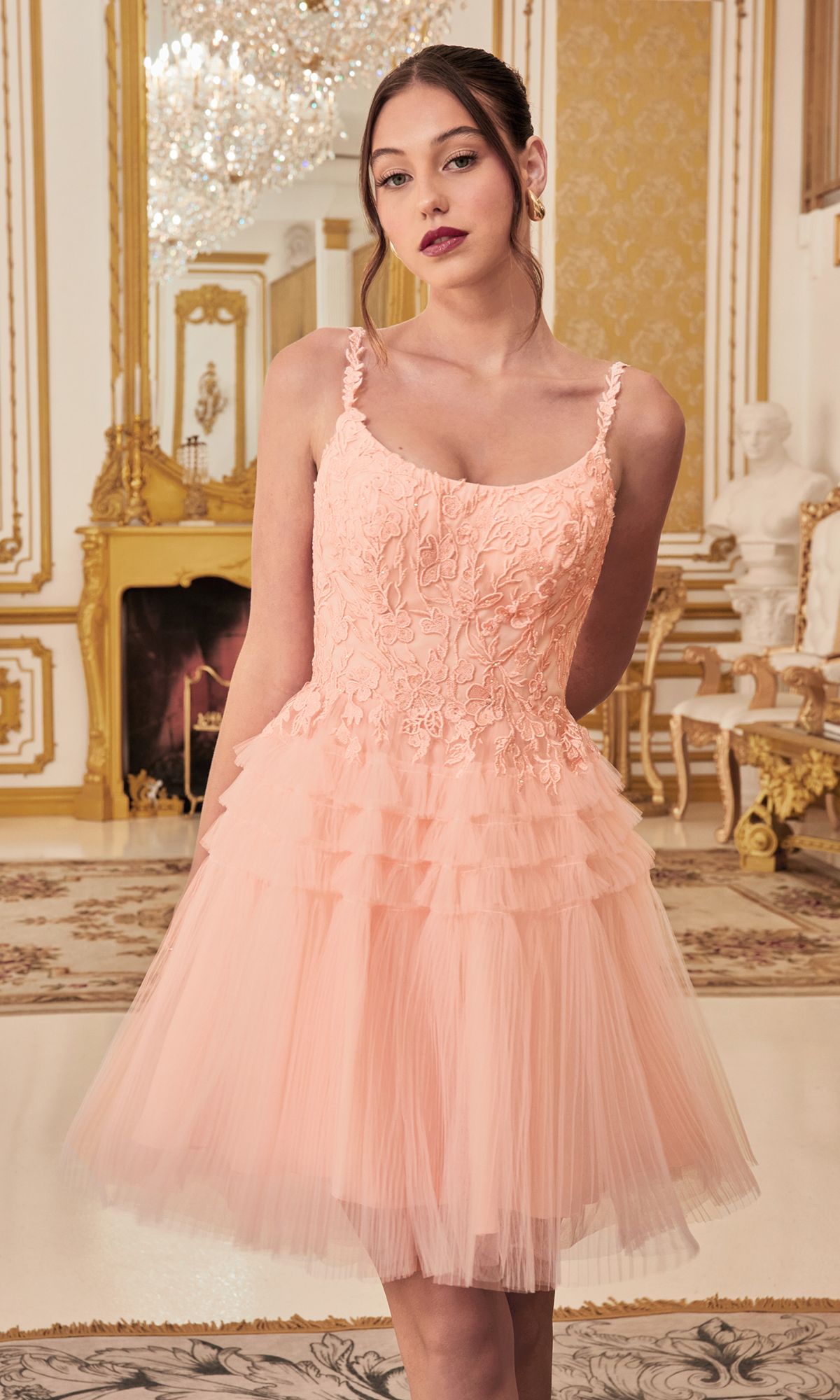 A woman stands indoors in an ornate, gold-accented room, wearing the Lace-Bodice Tiered-Skirt Short Prom Dress 9310 featuring a light pink lace bodice and a fitted, tiered tulle skirt.